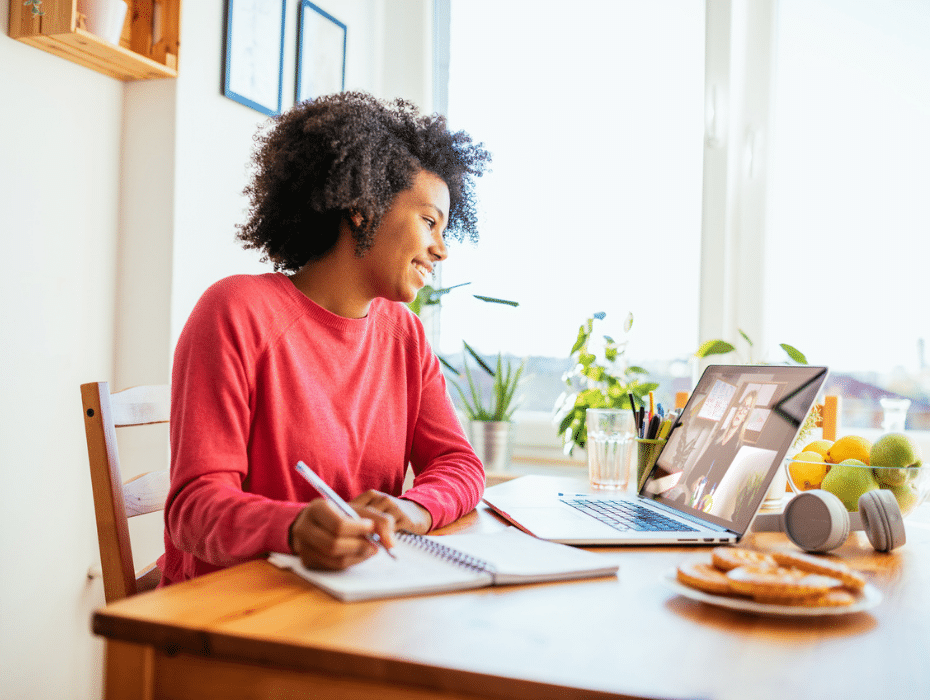 A woman sits at a table with a laptop and a notebook, focused on her work.