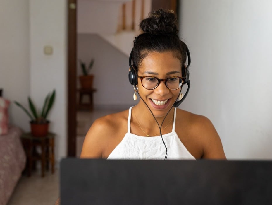 A smiling woman with glasses and a headset uses a laptop, engaged in her work or a virtual meeting.