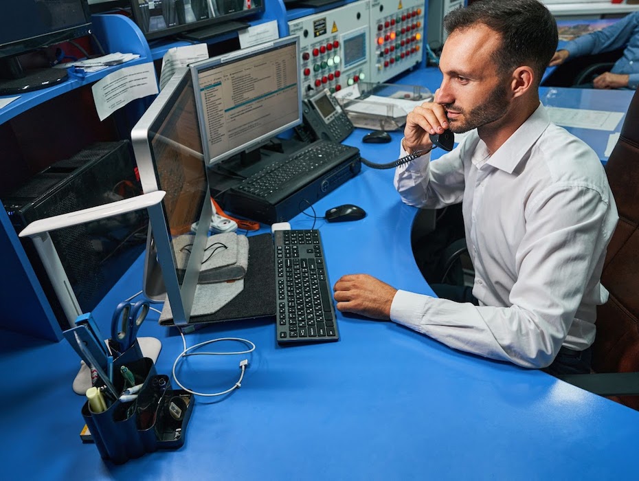 Caucasian male employee of a state and local government sitting at desk in open office in front of a PC while on a hardphone