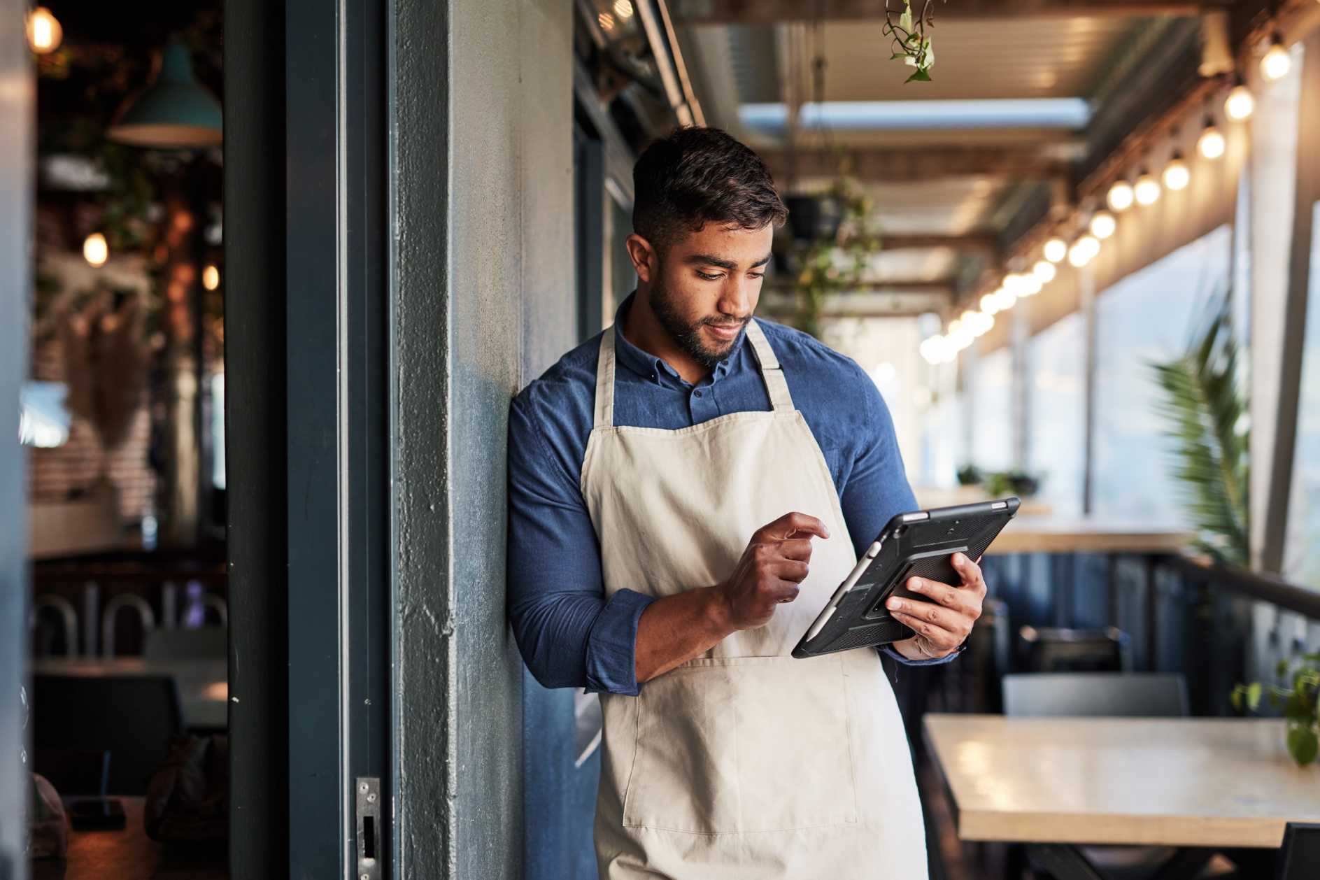 A man in an apron uses a tablet while standing in a restaurant, engaged in his work.